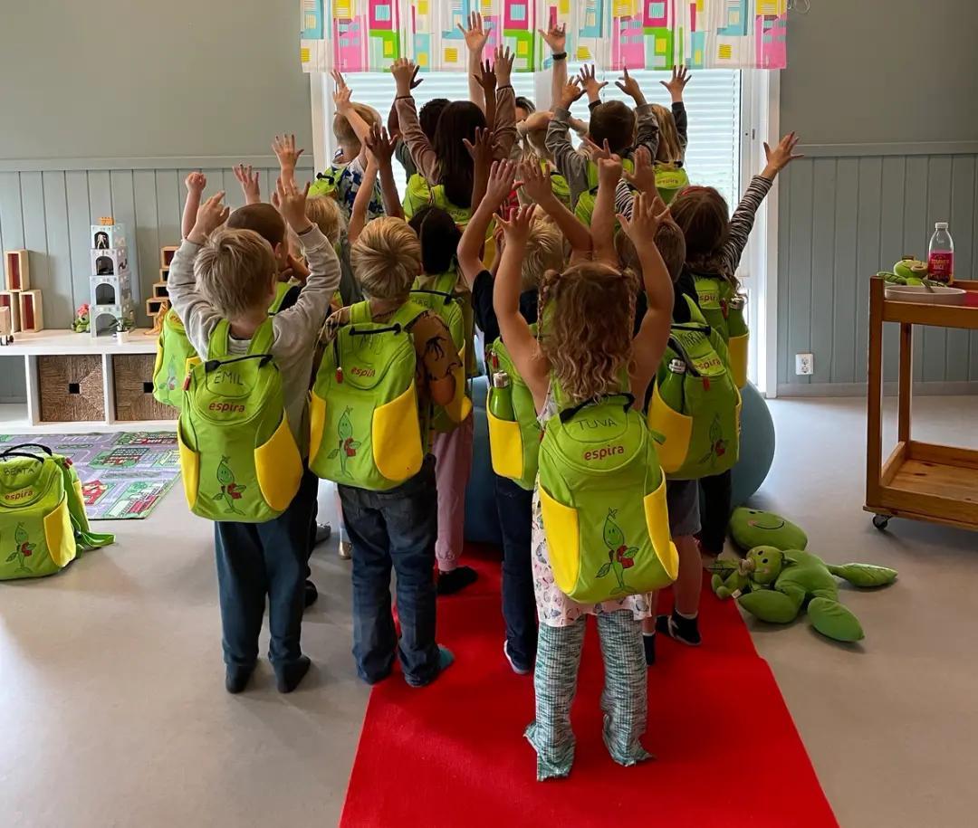 a group of children wearing green backpacks are standing in a room with their hands in the air .