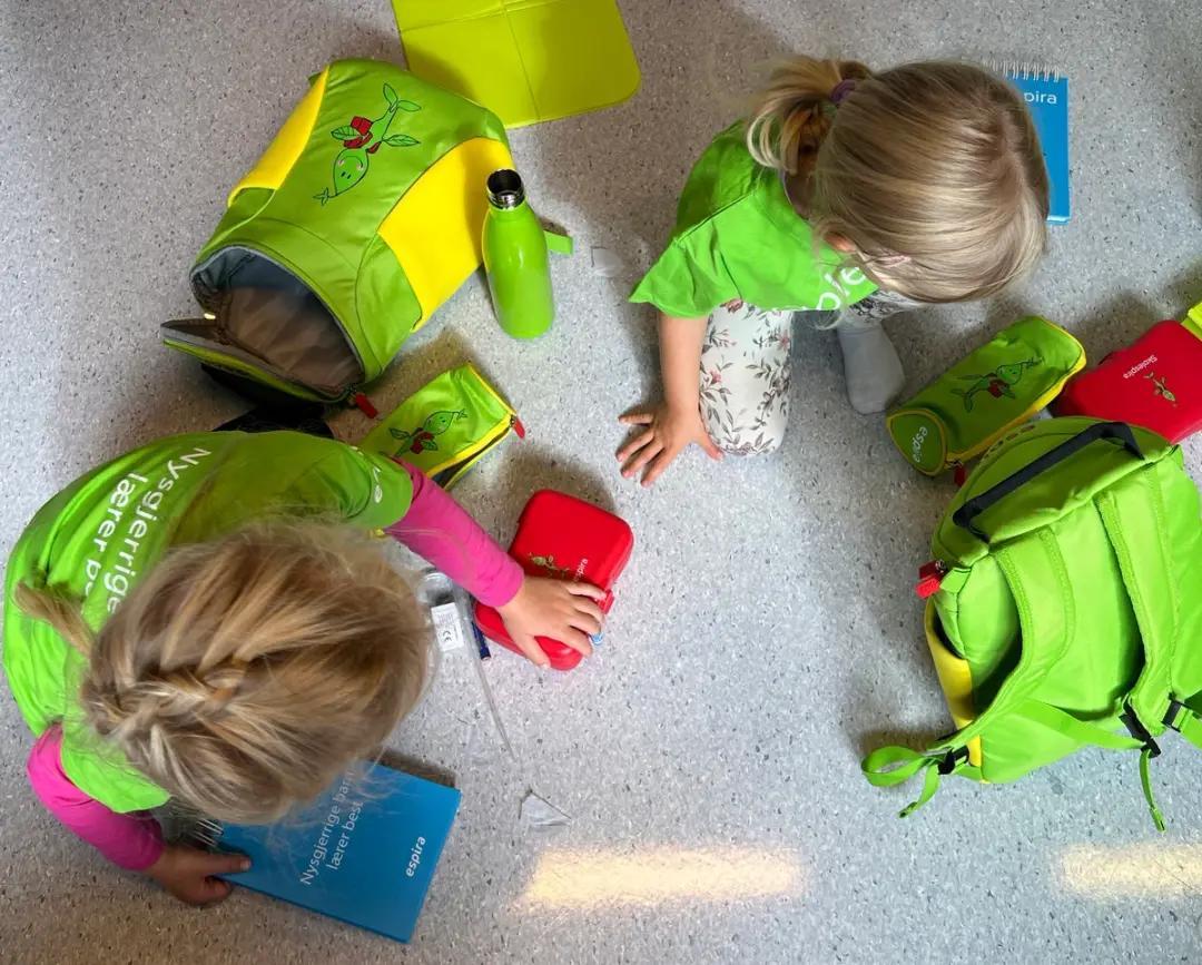 two little girls are sitting on the floor with their backpacks and lunch boxes .