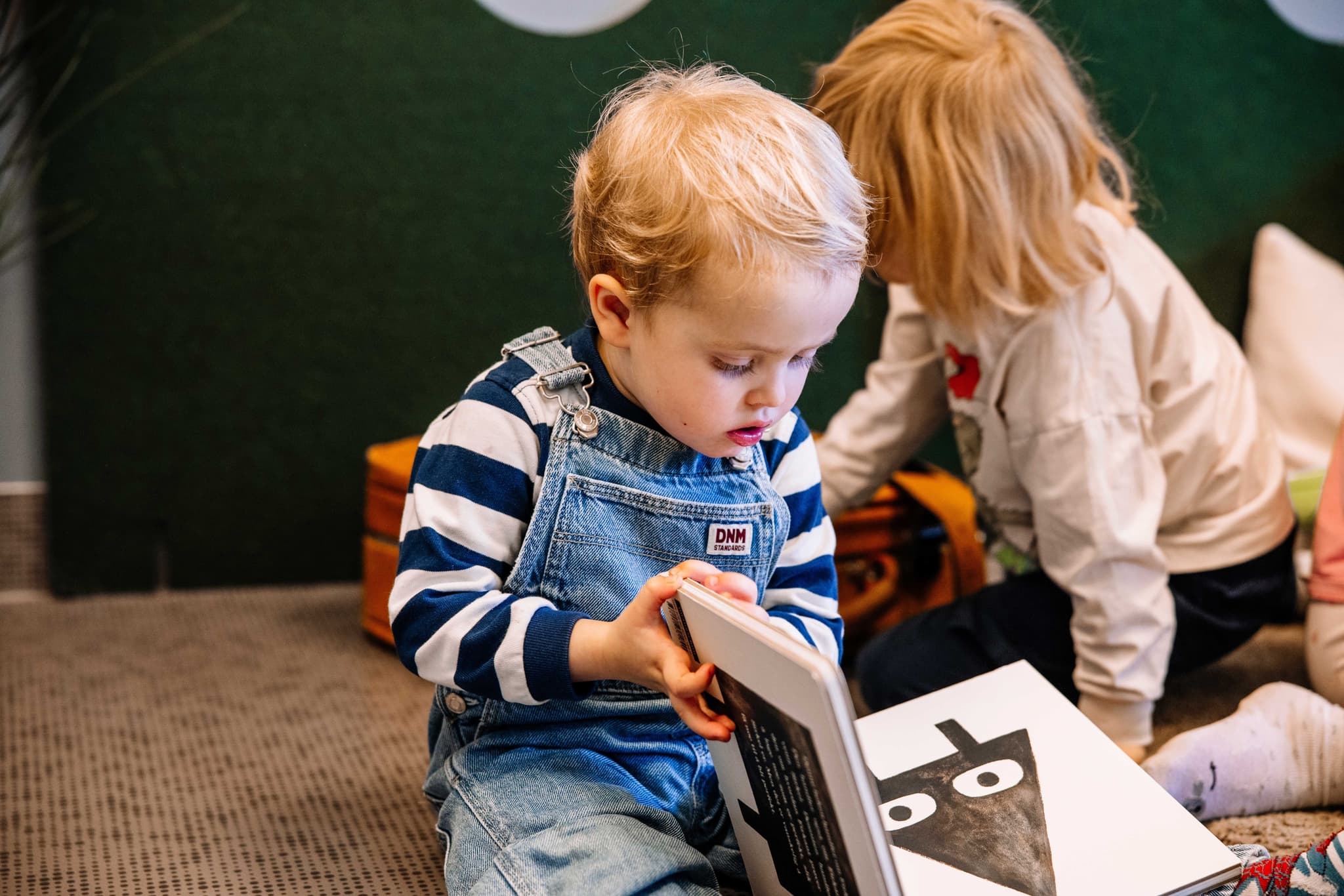 A young child in overalls sits on the floor, looking intently at a picture book.