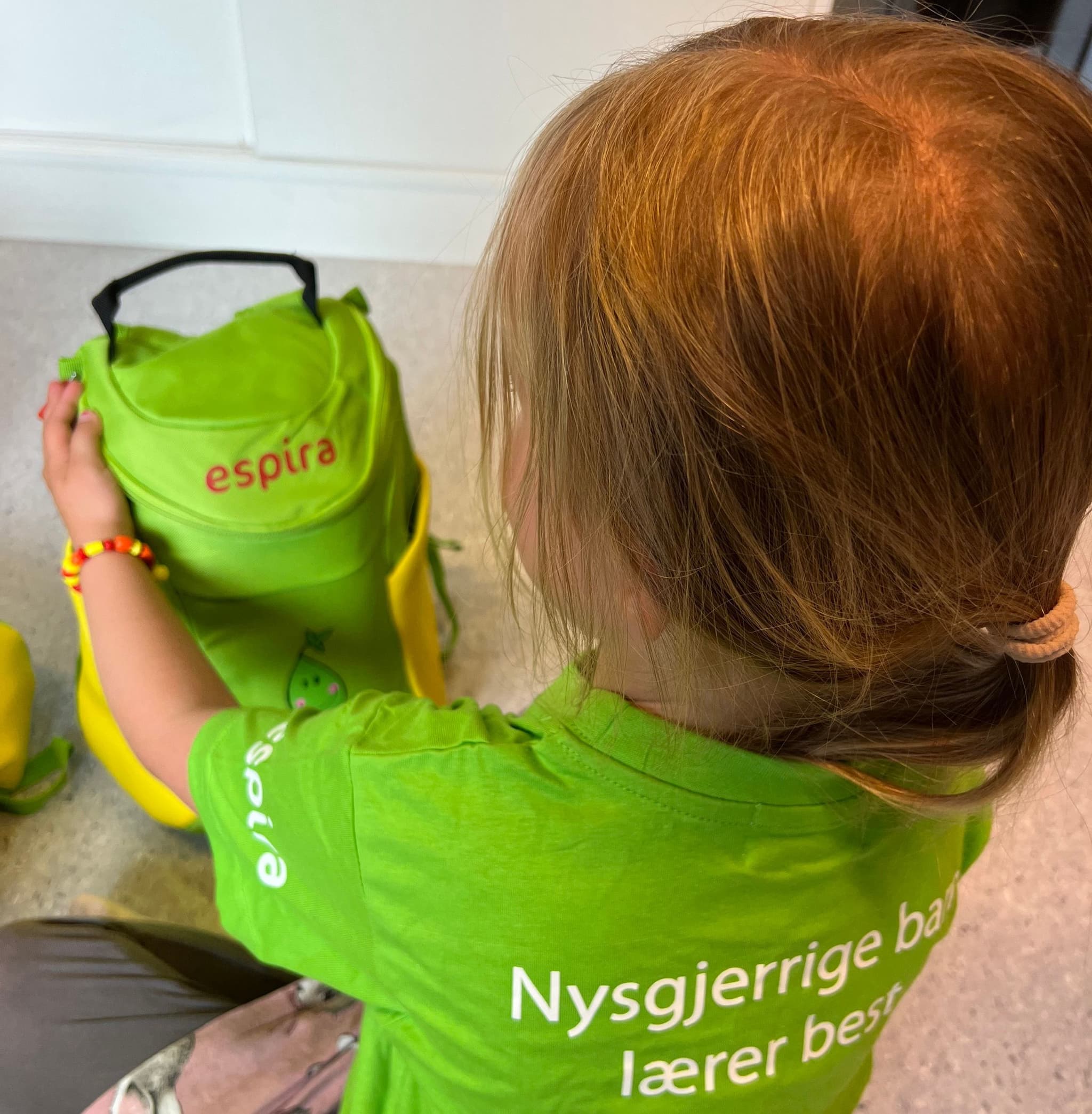 a little girl in a green shirt is holding a green backpack .