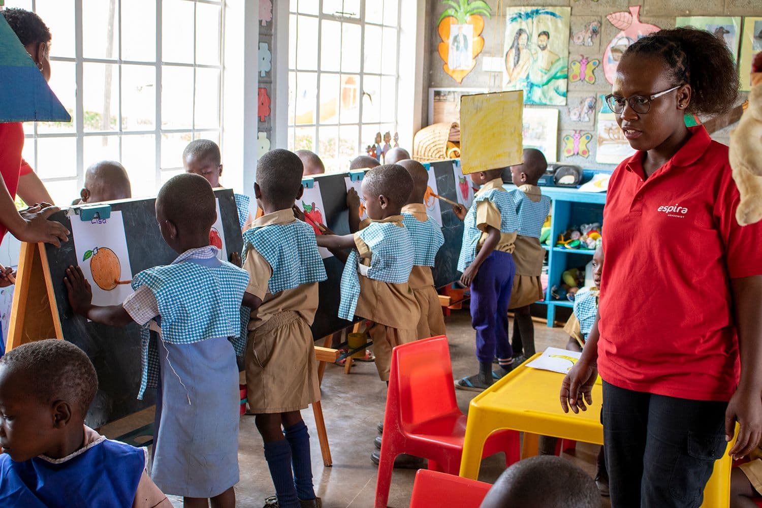 Young children draw on easels in a bright classroom, supervised by a teacher.