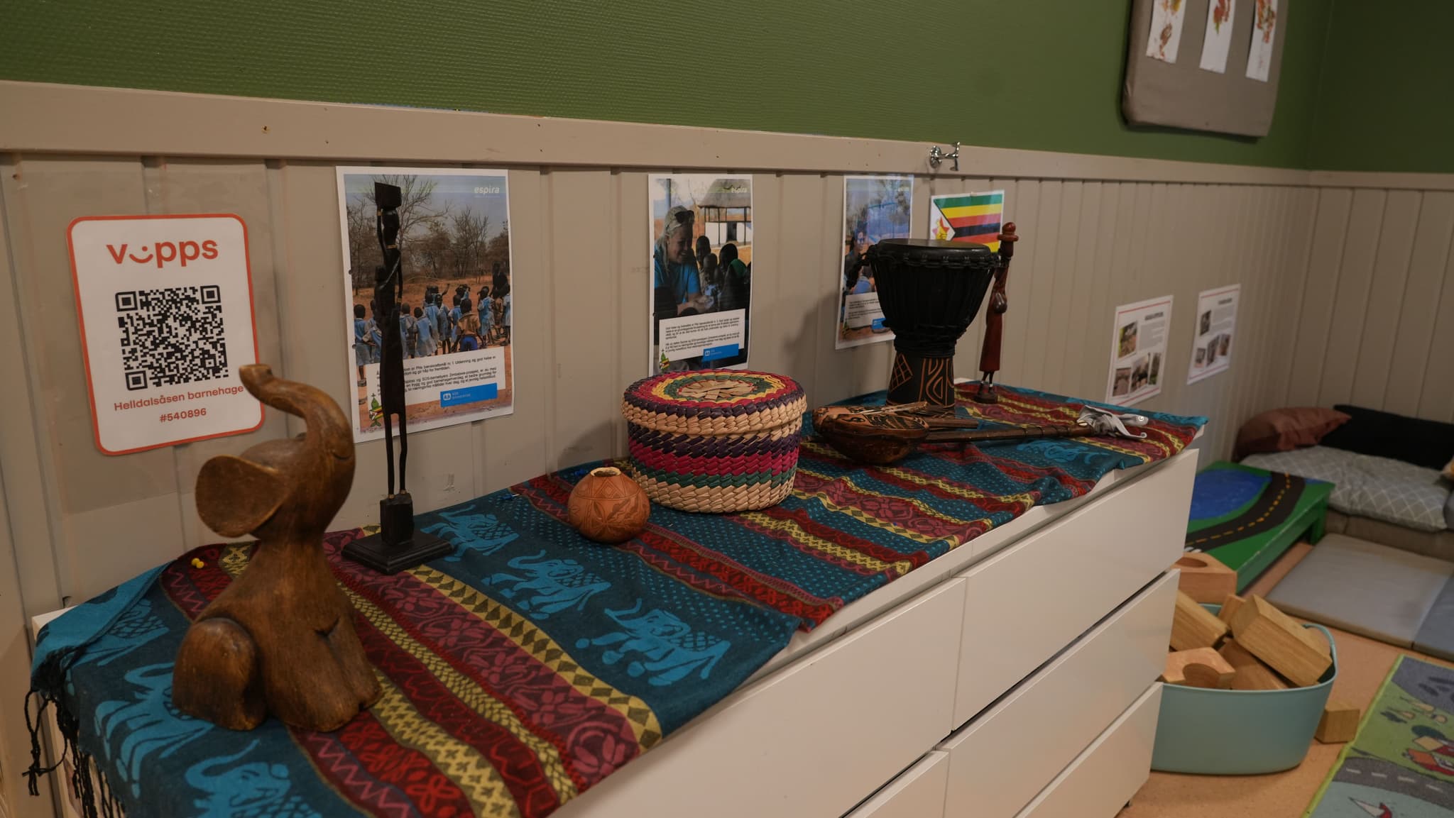 A dresser covered with patterned fabric displays a wooden elephant, a dark statue, a woven basket, and a djembe drum, with posters on the wall behind and a play area to the right.